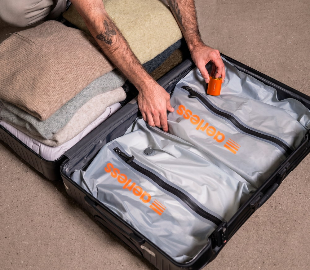 One open carry-on holding both states at once: a loose stack of folded knitwear filling the left half, and the same kind of clothing drawn down flat inside two Aerless bags on the right, with a hand setting the orange pump down beside them
